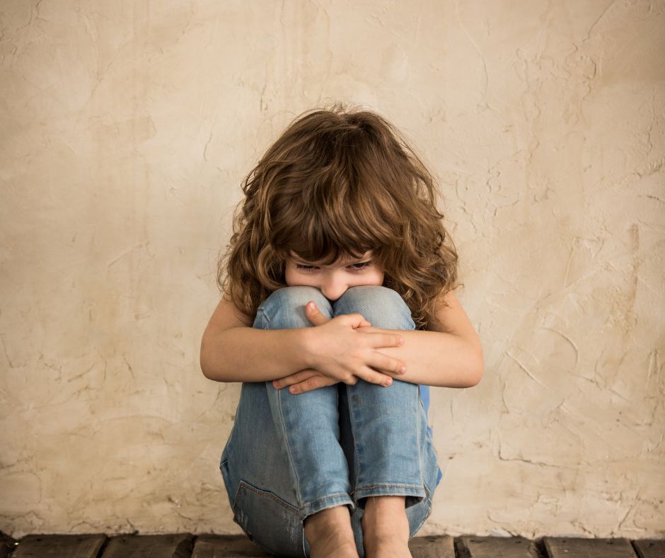 Ein Kind mit angezogenen Knien, um die es seine Arme gelegt hat. Sein Gesicht hat es zwischen den Knien versteckt.