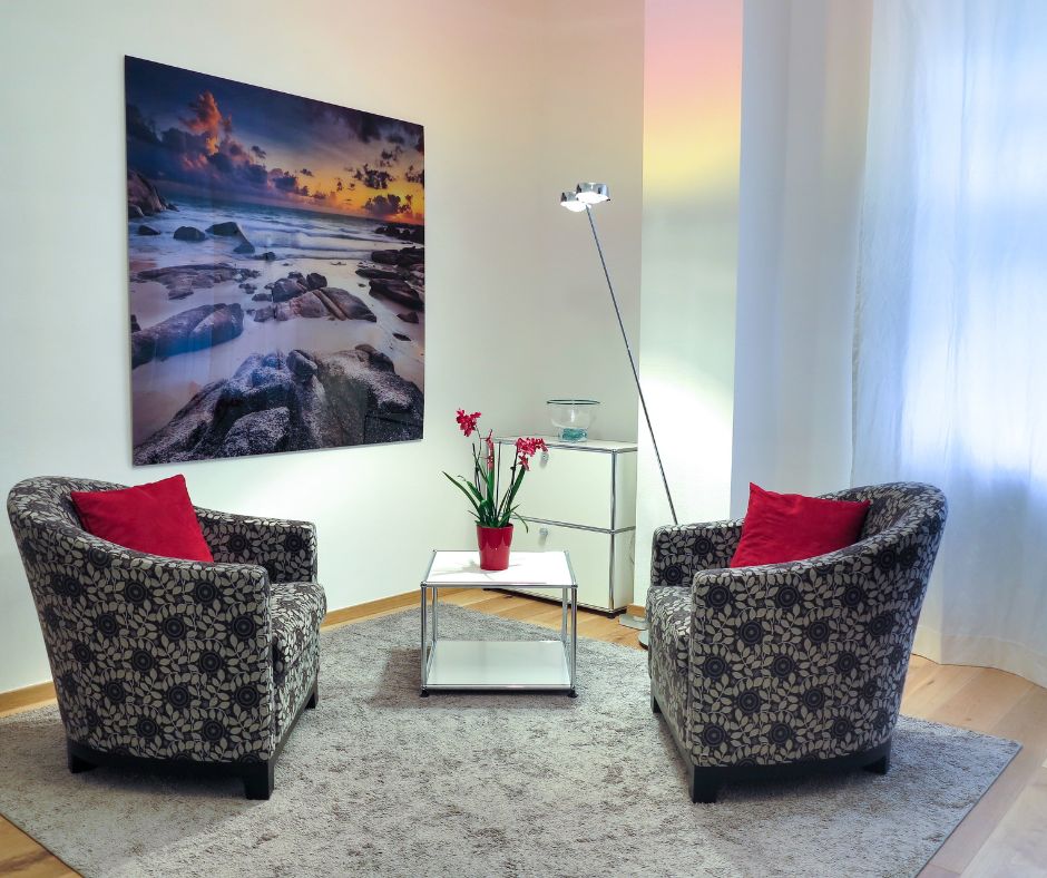 In einem Raum stehen sich zwei Sessel gegenüber. An der Wand hängt ein Bild. Etwas versetzt zwischen den Sesseln steht ein kleiner Tisch.